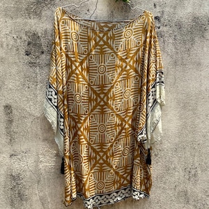 Puede incluir: Un caftán de color amarillo mostaza y blanco con un patrón geométrico repetitivo de círculos y líneas. La prenda tiene un escote barco ancho, mangas cortas estilo kimono y un dobladillo adornado con pequeñas borlas negras en el borde.
