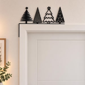 Op de afbeelding: Een zwarte metalen deurdecoratie met een rij van vier verschillende gestileerde kerstboom-silhouetten. Het ontwerp bevat bomen met horizontale lijnen, wervelende patronen, geschulpte randen met een ster en een herhalend boogmotief.