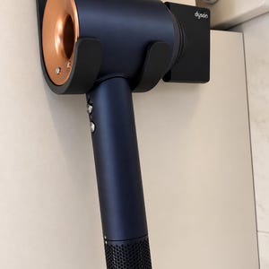 Op de afbeelding: Een donkerblauwe Dyson haardroger met een metallic koperkleurige ronde luchtuitlaat, gemonteerd in een zwarte wandhouder. Het apparaat heeft een lange handgreep met zilveren bedieningsknoppen, een geperforeerde onderkant en een zwart snoer.