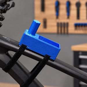 Könnte beinhalten: Eine leuchtend blaue, rechteckige, 3D-gedruckte Aufbewahrungshalterung, die am Oberrohr eines mattschwarzen Mountainbike-Rahmens befestigt ist. Die Halterung verfügt über einen zylindrischen Pfosten und wird mit zwei schwarzen Klettbändern am Fahrradrahmen befestigt.