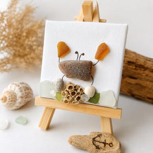 Könnte beinhalten: Eine kleine quadratische Leinwand auf einer Holzstaffelei mit einem Krabbenmotiv aus einem braunen Stein und zwei bernsteinfarbenen Meerglasstücken. Die Krabbe sitzt auf einer Ansammlung kleiner weißer Muscheln, grünem Meerglas und beigem Korallenstück.