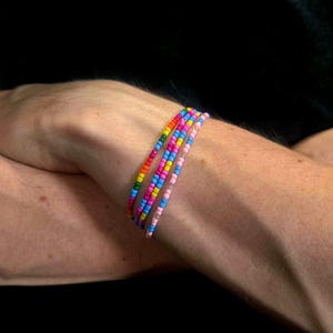 Könnte beinhalten: Drei dünne Armbänder aus kleinen, bunten Saatperlen. Ein Armband zeigt eine Regenbogenabfolge aus Rot, Orange, Gelb, Grün und Blau. Die anderen beiden Armbänder bestehen aus abwechselnden rosa, violetten und hellblauen Perlen.