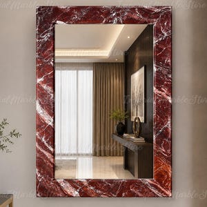 May include: A rectangular wall mirror featuring a thick, polished frame made of deep red marble with prominent white veining. The reflective glass surface displays a clear view of a room interior, including a console table and curtains.