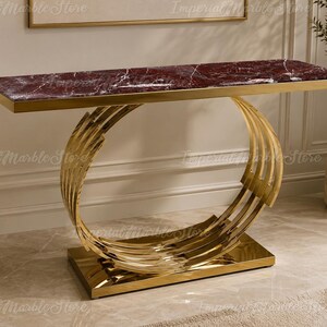 May include: A rectangular console table featuring a deep burgundy marble top with white veining. The base is constructed from polished gold-tone metal, consisting of multiple curved, circular rings that connect the tabletop to a rectangular base plate.