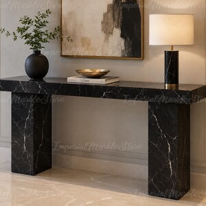 Può includere: Un tavolo console rettangolare in marmo nero con venature bianche, sostenuto da due spesse gambe a blocco. Sul tavolo si trovano un vaso nero con fogliame verde, un libro intitolato STONE con una ciotola dorata sopra e una lampada in marmo.