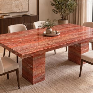 Puede incluir: Una mesa de comedor rectangular con una superficie de piedra travertino roja pulida con vetas naturales y una base de pedestal sólida y gruesa. La mesa está rodeada por seis sillas tapizadas en tela beige claro con patas de madera oscura.