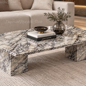 Puede incluir: Una mesa de centro rectangular con una superficie de mármol pulido en blanco y gris con vetas negras prominentes. La mesa se apoya sobre dos patas de bloque de mármol grueso a juego y sostiene un cuenco oscuro, dos libros y un florero.
