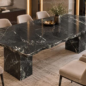 Puede incluir: Una gran mesa de comedor rectangular con una superficie de mármol negro pulido con vetas blancas y un borde natural. La mesa está sostenida por dos bases de pedestal gruesas de mármol negro a juego y rodeada por seis sillas tapizadas en beige.