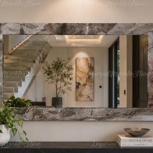 May include: A rectangular wall mirror featuring a wide frame made of grey and white veined marble. The mirror reflects an interior space with a staircase, a potted plant, and wall art. The text Imperial Marble Store is visible on the surface.