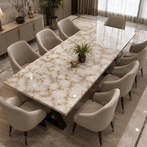 May include: A rectangular dining table with a white and gold veined marble top and a black metal base, surrounded by eight beige upholstered chairs with vertical stitching and dark legs. A small green plant sits in the center of the table.