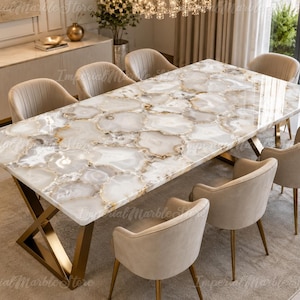 May include: A rectangular dining table featuring a polished white and beige agate gemstone slab tabletop with a natural crystalline pattern, supported by a gold-finished metal X-shaped base, surrounded by six beige upholstered chairs.