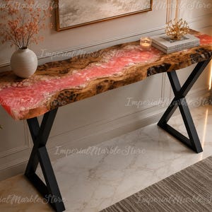 Può includere: Un tavolo console caratterizzato da un piano in legno naturale con bordo irregolare e un fiume centrale in resina rosa traslucida. Il tavolo è sostenuto da due gambe in metallo nero a forma di X. Il piano ospita un vaso bianco, una candela e libri.