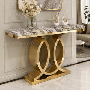 May include: A rectangular console table featuring a white and grey marble top and a polished gold metal base with two interlocking circular supports. The table is adorned with a clear glass vase containing white flowers and two white swan figurines.