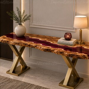 May include: A console table featuring a live-edge wood slab with a central deep red resin river inlay. The table is supported by two gold-colored metal X-shaped legs with rectangular bases. A white vase, a candle holder, and books sit on top.