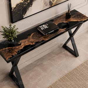 May include: A rectangular console table featuring a live-edge wood slab centrepiece encased in a glossy black resin surface. The table is supported by a black metal X-shaped frame base. A small green plant and a Tom Ford book sit on top.