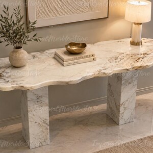 Peut inclure: Une console en marbre blanc avec un bord organique naturel et irrégulier, soutenue par deux pieds rectangulaires. La surface présente des veines grises. Un vase en céramique beige avec de la verdure, deux livres et un bol doré sont posés dessus.