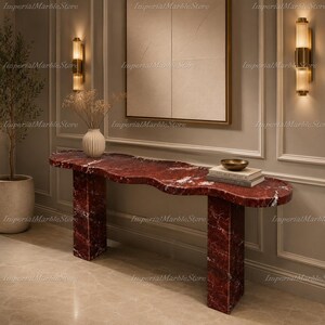 May include: A console table crafted from deep red marble with prominent white veining, featuring a thick, organic-shaped top supported by two rectangular slab legs. The surface holds a small stack of books and a shallow metallic bowl.