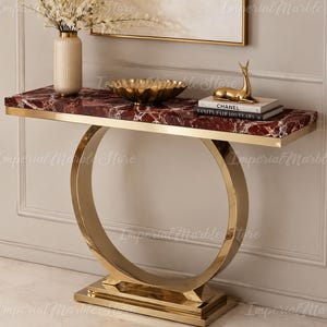 May include: A console table featuring a rectangular deep red marble top with white veining, supported by a circular gold-toned metal base. The table holds a ribbed cream vase, a gold scalloped bowl, a gold deer figurine, and two stacked books.