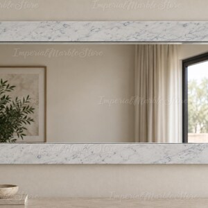 May include: A rectangular wall mirror featuring a wide, flat frame crafted from white marble with grey veining. The mirror surface reflects a room interior with a framed picture, a plant, and a window with curtains, set against a neutral wall.