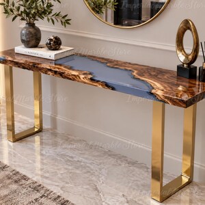 Pode incluir: Uma mesa consola retangular com um tampo de madeira de bordo natural e uma incrustação central de resina azul. A mesa é suportada por duas pernas em forma de U de metal dourado polido. A superfície é lisa e brilhante com veios.
