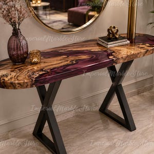 May include: An oval console table featuring a live edge wood slab with a deep purple resin inlay, supported by a black metal X-shaped base. The tabletop displays natural wood grain patterns and a glossy finish, holding a purple glass vase and books.