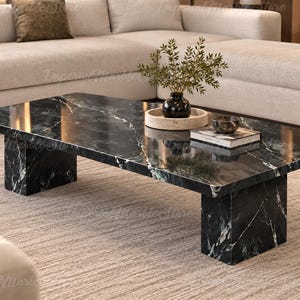 Peut inclure: Une table basse rectangulaire en marbre noir poli avec des veines blanches marquées. La table présente un plateau épais et plat soutenu par deux pieds larges et massifs. La surface est lisse et réfléchissante, révélant des motifs en pierre naturelle.