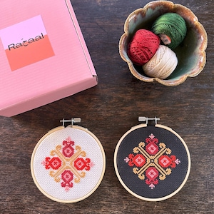 Pode incluir: Dois bastidores de bordado em ponto cruz com desenhos geométricos, um em tecido branco e outro em preto. Uma caixa rosa com o texto "Razaal" e uma tigela com fios de bordar vermelhos, verdes e creme também são visíveis.