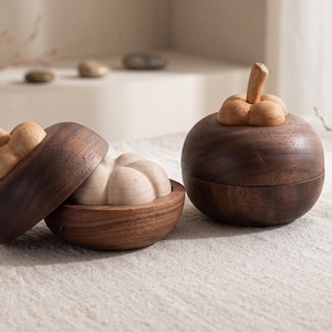 Puede incluir: Dos recipientes de madera con forma de mangostán, elaborados en madera de color marrón oscuro con tallos de madera clara. Un recipiente está cerrado, mientras que el otro está abierto, revelando una pieza interior segmentada de color claro.