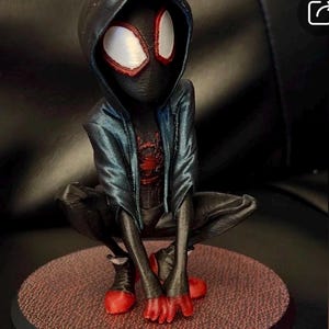 May include: A 3D printed figurine of a character wearing a black hooded jacket over a black suit with a red spider emblem on the chest. The character has large white eyes with red outlines, red fingertips, and red shoes, posed in a crouched position.