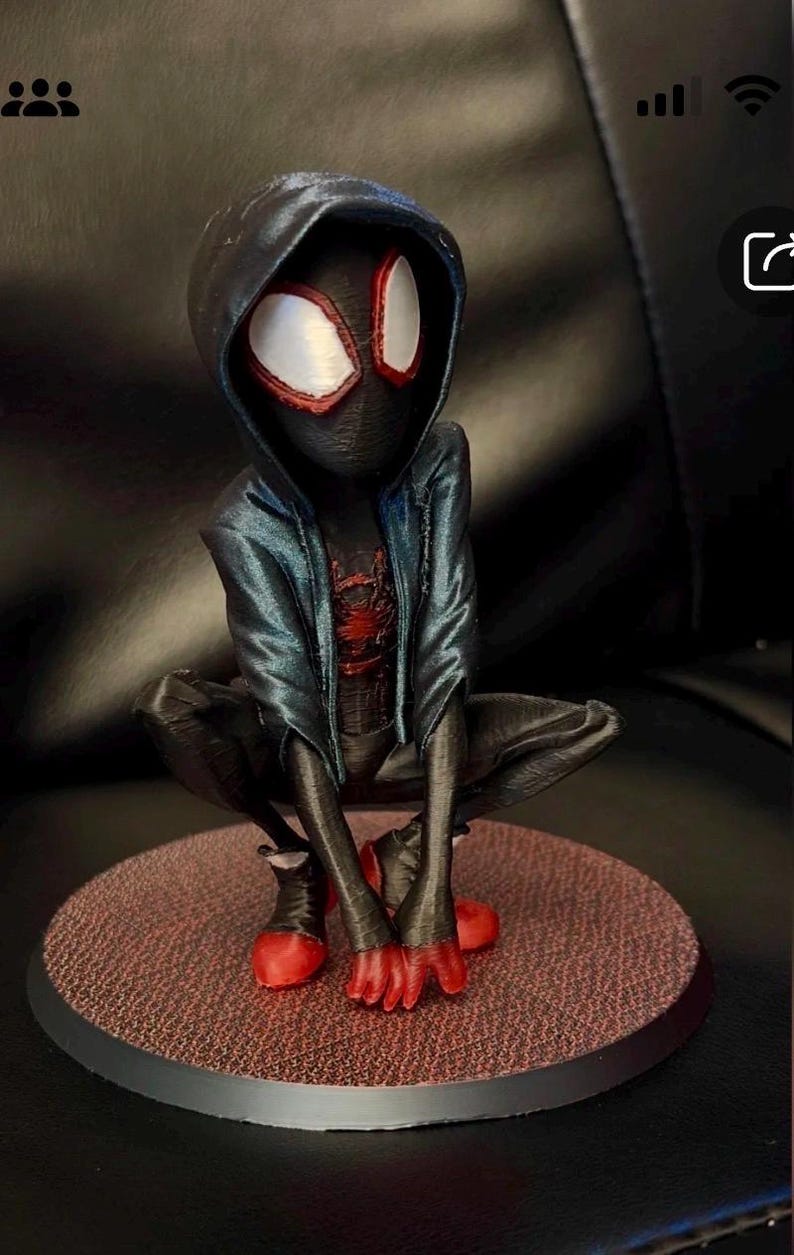 May include: A 3D printed figurine of a character wearing a black hooded jacket over a black suit with a red spider emblem on the chest. The character has large white eyes with red outlines, red fingertips, and red shoes, posed in a crouched position.