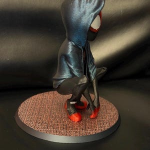 May include: A figurine of a character in a crouching pose, wearing a dark blue hooded jacket over a black suit with red accents on the shoes and hands. The figure is positioned on a circular base with a textured, reddish-brown grid pattern.