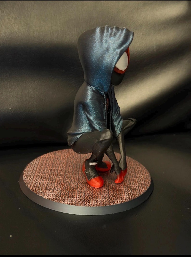 May include: A figurine of a character in a crouching pose, wearing a dark blue hooded jacket over a black suit with red accents on the shoes and hands. The figure is positioned on a circular base with a textured, reddish-brown grid pattern.