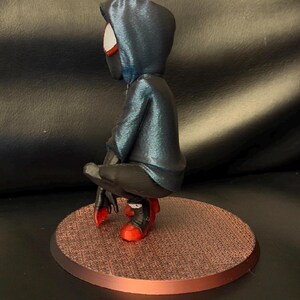 May include: A stylized figurine of a character in a crouching pose, wearing a dark blue hooded jacket, black pants, and red and black sneakers. The figure is positioned on a circular, textured copper-colored base, viewed from the side profile.