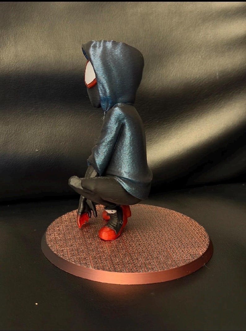 May include: A stylized figurine of a character in a crouching pose, wearing a dark blue hooded jacket, black pants, and red and black sneakers. The figure is positioned on a circular, textured copper-colored base, viewed from the side profile.