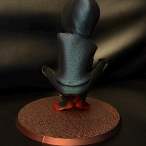 May include: A figurine of a hooded character in a dark blue, shimmering finish, shown from the back. The figure is in a crouched position, wearing bright red footwear, and is mounted on a circular, textured, copper-colored base.