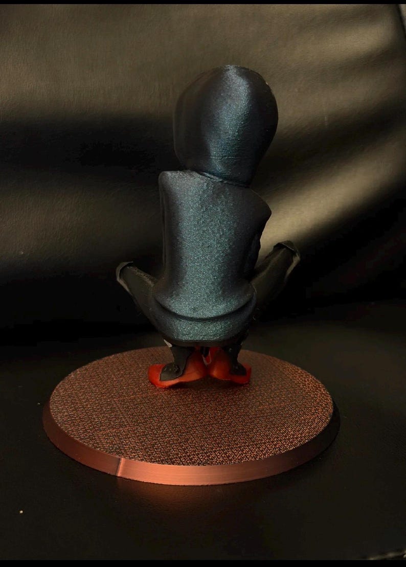 May include: A figurine of a hooded character in a dark blue, shimmering finish, shown from the back. The figure is in a crouched position, wearing bright red footwear, and is mounted on a circular, textured, copper-colored base.