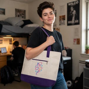 Könnte beinhalten: Eine cremefarbene Canvas-Tragetasche mit einem dunkelvioletten Boden und passenden violetten Schulterriemen. Die Tasche verfügt über eine Reißverschlusstasche auf der Vorderseite mit einer bunten, abstrakten Linienzeichnung in Pink, Blau und Gelb.