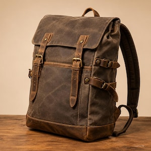 Peut inclure: Un sac à dos en toile cirée marron foncé doté d'un rabat supérieur avec deux sangles en cuir verticales, des boucles en laiton et des sangles de compression latérales. La base est renforcée en cuir marron, avec une poignée et des bretelles.