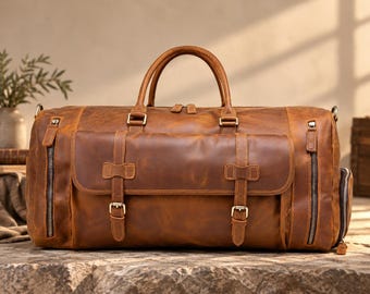 May include: A tan leather duffel bag featuring two top handles, a large front pocket with two buckle straps, and two vertical zippered side pockets. The bag has a smooth, matte finish with visible stitching and metallic hardware accents throughout.