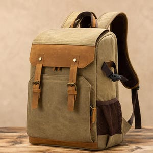 Könnte beinhalten: Ein khakifarbener Rucksack aus Segeltuch mit braunen Lederakzenten, einer oberen Klappe mit zwei Schnallenriemen, einer Vordertasche und einer dunkelbraunen Netzseitentasche. Der Rucksack verfügt über verstellbare Schultergurte und Metallbeschläge.