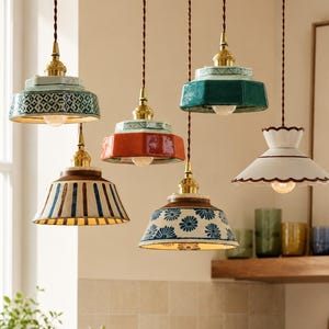 Può includere: Una collezione di sei lampade a sospensione con paralumi in ceramica di varie forme e colori, tra cui verde acqua, arancione, bianco e motivi blu e bianchi, sospese tramite cavi intrecciati marroni con supporti color ottone.