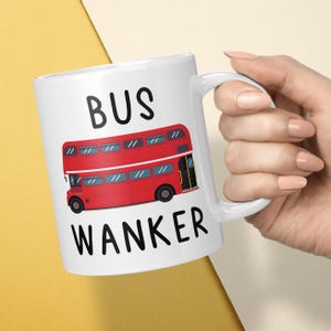 May include: A white ceramic mug featuring a graphic illustration of a red double-decker bus centered between the black text BUS and WANKER. The mug has a standard handle on the right side and a smooth, glossy finish throughout the exterior surface.