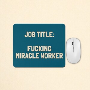 May include: A rectangular teal mouse pad featuring the text "JOB TITLE: FUCKING MIRACLE WORKER" in a bold, cream-coloured sans-serif font. A white computer mouse with a scroll wheel is positioned to the right of the mouse pad on the surface.