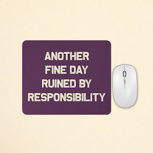 May include: A rectangular dark purple mouse mat featuring the white distressed-style text ANOTHER FINE DAY RUINED BY RESPONSIBILITY, positioned next to a white computer mouse with a scroll wheel and two buttons on a light beige surface.