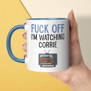 May include: A white ceramic mug with a blue handle and interior rim. The front features the blue text FUCK OFF and black text I'M WATCHING CORRIE above a graphic of a vintage television set displaying a brick building labeled CORONATION ST.