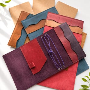 May include: A collection of rectangular leather notebook covers in various colors including deep purple, bright red, blue, and tan. Each cover features internal pockets with wavy edges and a central purple elastic cord system for holding inserts.