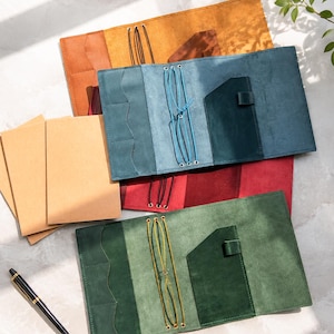 May include: A collection of four leather notebook covers in mustard yellow, blue, red, and green. Each cover features internal pockets, elastic bands for securing inserts, and a pen loop. Three plain tan paper notebooks and a black fountain pen are included.