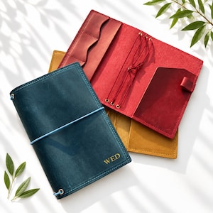 May include: A dark blue leather journal cover with the letters WED embossed in gold on the front, secured by a light blue elastic band. Beneath it lie an open red leather cover with internal pockets and elastic cords, and a yellow leather cover.