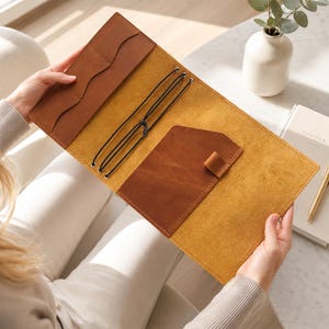 May include: A tan leather journal cover featuring a mustard yellow interior, multiple card slots, a pen loop, and black elastic cords for securing inserts. The cover is open, displaying its internal pockets and binding system for holding notebooks.
