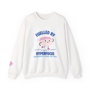 Puede incluir: Una sudadera blanca de manga larga con un gráfico de un cerebro de dibujos animados con una cinta rosa y zapatillas de correr. El texto dice "FUELLED BY HYPERFOCUS NEUROSPICY MUM EDITION" en azul, con texto adicional en la manga.